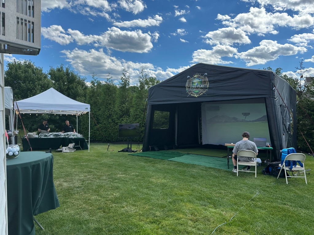 Branded golf simulator tent on a sunny lawn with a catering table and attendants.